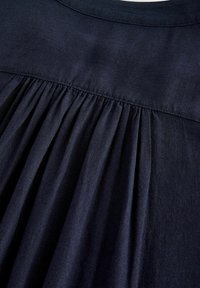 Tamno mornarsko plava tkanina s mekom teksturom, koja se ističe nabranim detaljima na leđima i jednostavnim okruglim izrezom, naglašavajući svoj drapirani dizajn.