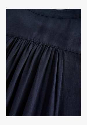 Donker marineblauwe stof met een zachte textuur, voorzien van plooien aan de achterkant en een eenvoudige ronde halslijn, die het gedrapeerde ontwerp benadrukt.