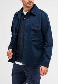 Marineblaue Jacke mit langen Ärmeln, zwei Brusttaschen, Knopfverschluss und einem leicht strukturierten Stoff, über einem dunklen Pullover getragen.