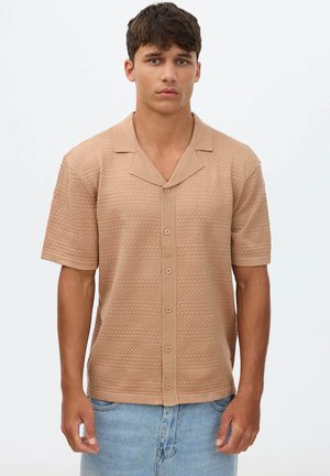 Joven con el pelo corto y castaño que lleva una camisa beige texturizada de manga corta abotonada y vaqueros azul claro, de pie frente a un fondo blanco.
