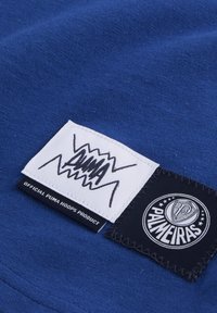Blåt stof med to vævede labels: en hvid med sorte grafikker, der læser "PUMA," og en anden sort med et rundt logo, der siger "PALMEIRAS."