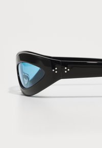 Lunettes de soleil noires avec un design élégant et anguleux et des verres bleus. Présentent de petits accents métalliques sur la monture. Texture lisse et brillante.