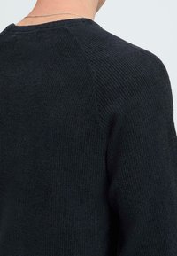 Zwarte gebreide trui met raglanmouwen; toont de nek en schouder om details over het materiaal en de constructie te laten zien.