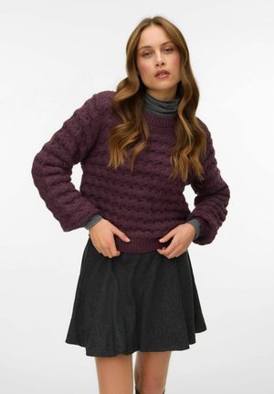 Strikket sweater i dyb lilla med struktureret bølget mønster, rund halsudskæring og lange ærmer, parret med en flagrende mørkegrå nederdel.