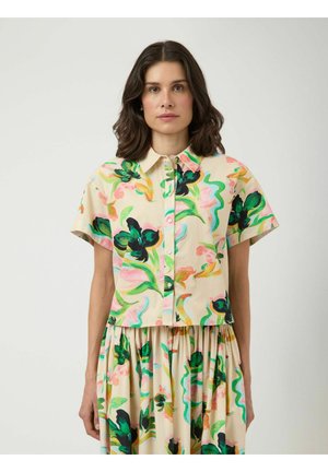 Kvinde står vendt fremad iført en kortærmet skjorte med knapper og et skørt med et farverigt blomstermønster på en lys beige baggrund.