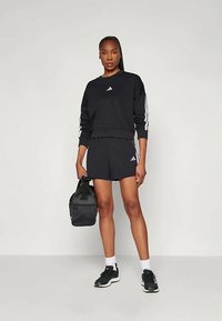 Črna športna trenirka z belimi črtami, ujemajoče črne kratke hlače in črne superge. Črna torba je v eni roki.