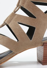 Sandale en daim beige à bouts ouverts avec un design découpé, doublure noire et un talon en bois épais. Semelle plate et finition texturée.