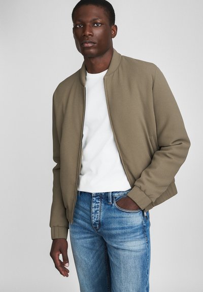 Chaqueta bomber caqui con puños acanalados, llevada sobre una camiseta blanca, combinada con jeans de mezclilla azules. Textura de tela suave, diseño minimalista.