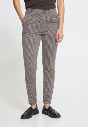 Persoon draagt een slim-fit, hoog getailleerde broek met diagonale strepen en zwarte leren loafers, staand tegen een effen lichte achtergrond.