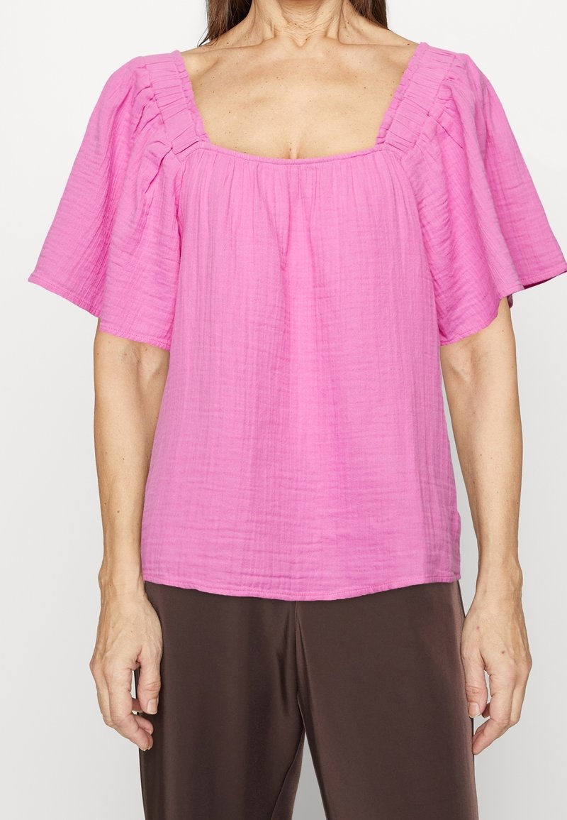 Haut rose à encolure carrée en tissu léger avec manches courtes bouffantes et motif texturé. Associé à un pantalon marron.