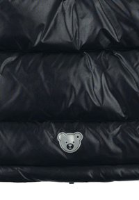 Tessuto imbottito nero con trapuntatura orizzontale e un piccolo logo riflettente di un orsetto grigio posizionato al centro verso il basso.
