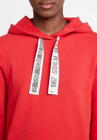 Sudadera roja con capucha y bolsillo canguro, fabricada en tejido texturizado, con cordones blancos que presentan un texto en patrón negro. Vista en primer plano del escote.