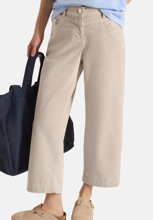 Beżowe spodnie z szerokimi nogawkami noszone z jasnoniebieską koszulą, beżowymi butami wsuwanymi i granatową torbą trzymaną w lewej ręce.