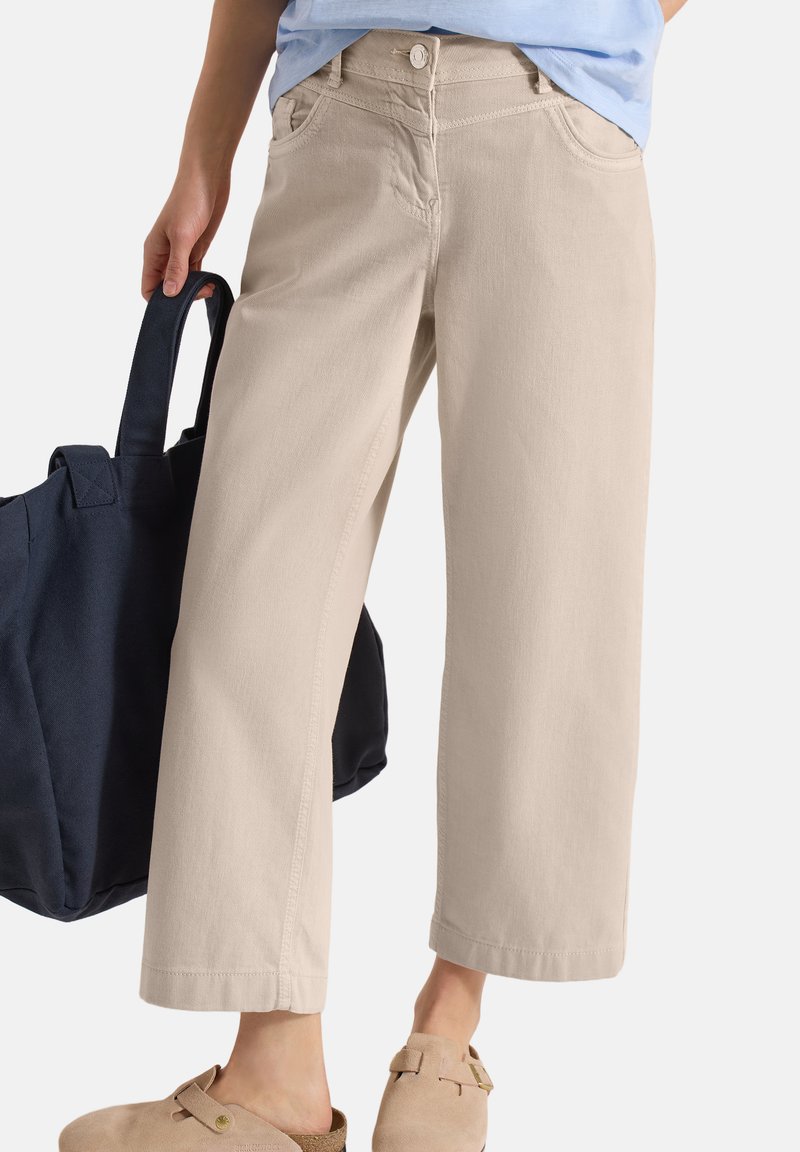 Beige bukser med vide ben båret med en lyseblå skjorte, beige slip-on sko og en mørkeblå taske holdt i venstre hånd.