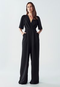 Ženska z rokami v žepih, oblečena v črno širokohlavo kombinezon z rokavi do komolcev in globokim V-izrezom.