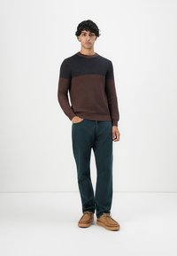 Un jeune homme debout avec une main dans la poche, portant un pulls côtelé marron et noir, un pantalon sombre et des chaussures en cuir tan à lacets, sur un fond blanc uni.