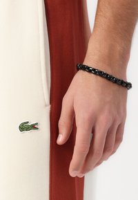 Rdeče in smetana jogger hlače z zeleno krokodilovo logotipno podrobnostjo, v paru z teksturiranim črnim verižnim zapestnikom, roka naslonjena na tkanino.