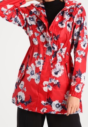 Femme porte un imperméable à capuche rouge à fleurs blanches et bleu marine, taille cintrée et poignets élastiques sur des vêtements noirs.
