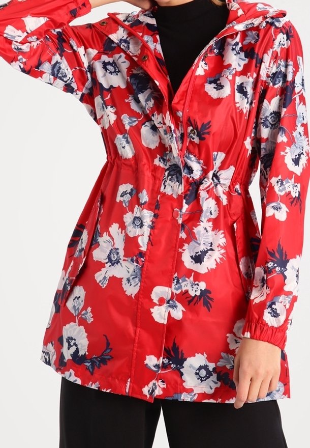 Femme porte un imperméable à capuche rouge à fleurs blanches et bleu marine, taille cintrée et poignets élastiques sur des vêtements noirs.