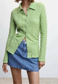 Chemise verte texturée à manches longues avec boutons et col, associée à une mini-jupe en denim bleu. Présente des boutons visibles et une coupe décontractée.