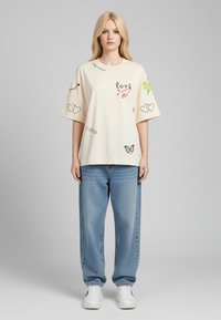 T-shirt surdimensionné couleur crème avec des impressions graphiques comprenant des cœurs, des fleurs et des papillons. Associé à un jean bleu clair ample et des baskets blanches.