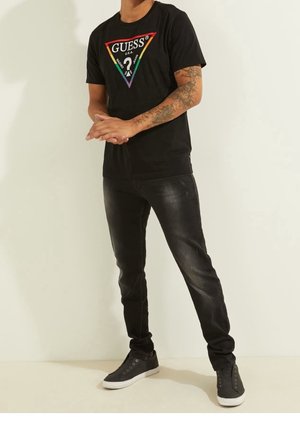 Mann trägt schwarzes Guess T-Shirt mit Regenbogen-Dreieck-Logo, schwarze Jeans und schwarze Sneakers, steht mit vor den Händen verschränkten Händen.