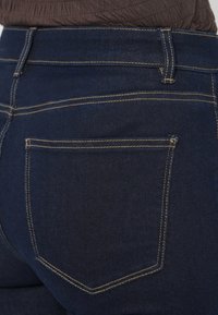 Σκούρο μπλε τζιν με ομαλή υφή, με αντίθετη κίτρινη ραφή και πίσω τσέπη με ενισχυμένη γωνία.