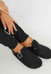 Zuecos de ante negro con una hebilla plateada cuadrada y una correa segura, con punta redondeada y suela texturizada, llevados con jeans negros ajustados.