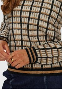 Pull en tricot avec un motif en grille beige, marron et noir. Comprend des poignets et un ourlet côtelés avec des détails rayés. La texture de la laine est visible.