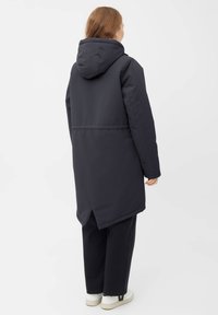 Dlouhý tmavě šedý kabát s kapucí, s volným střihem, hladkým povrchem a detailem rozparku na zadní straně. Spárován s tmavými kalhotami a bílými teniskami.