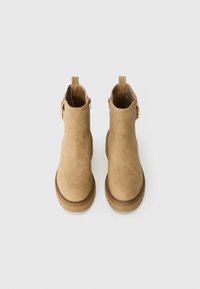 Bottines en daim beige avec un bout rond et une semelle en caoutchouc épaisse. Présentent des accents de fermeture éclair et une languette au niveau du talon.