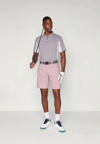 Άνδρας που φοράει γκρί πόλο μπλούζα γκολφ, ροζ σορτς, λευκό γάντι γκολφ και λευκά παπούτσια, κρατώντας ένα μπαστούνι γκολφ στον ώμο του σε μια ουδέτερη σκηνή.