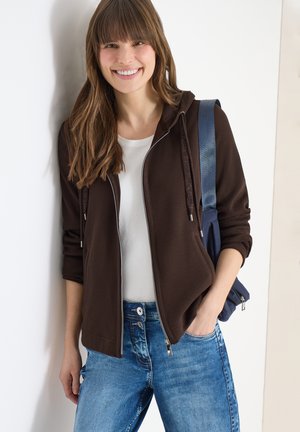 Femme souriante aux longs cheveux bruns portant un sweat à capuche marron zippé, une chemise blanche, un jean bleu et un sac à dos bleu marine, debout contre un mur blanc.