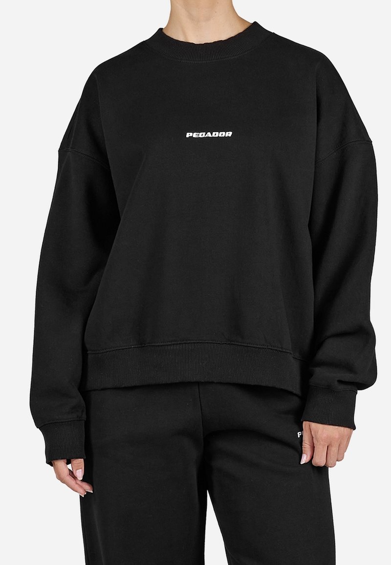 Personne portant un sweat-shirt noir à manches longues avec le texte « PEGADOR » centré sur la poitrine et un pantalon noir assorti.