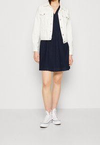 Vit denimjacka över en marinblå klänning med ögla-textur, tillsammans med vita höga sneakers. Enkel, avslappnad outfit med lager-på-lager-element.