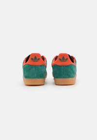Green suede sneakers with orange accents and black lining. Gum sole and adidas logo on the heel. Rounded shape with cushioned design.
