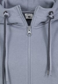 Hellgrauer Reißverschluss-Hoodie aus weichem Material, mit einem metallischen Reißverschluss, verstellbaren grauen Kordeln und einem Etikett, das die Größe M angibt.