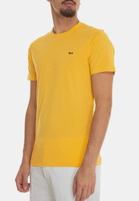 T-shirt giallo a maniche corte in cotone con scollatura rotonda e un piccolo logo di un cane nero sul petto. Tessuto liscio, vestibilità casual.