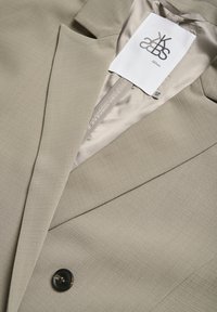 Beiger Blazer aus strukturiertem Stoff, mit Reverskragen, einem einzigen Knopfverschluss und einem kontrastierenden glatten Futter. Markenlabel sichtbar.