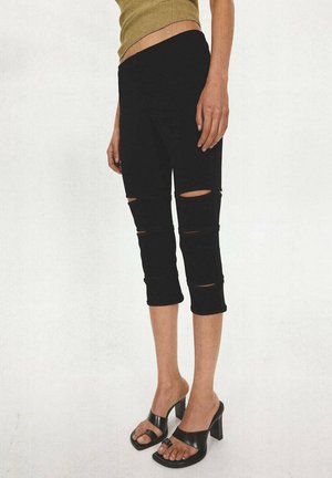Persoon draagt zwarte gescheurde cropped broek en zwarte sandalen met hak, met een deel van een groen mouwloos topje zichtbaar.