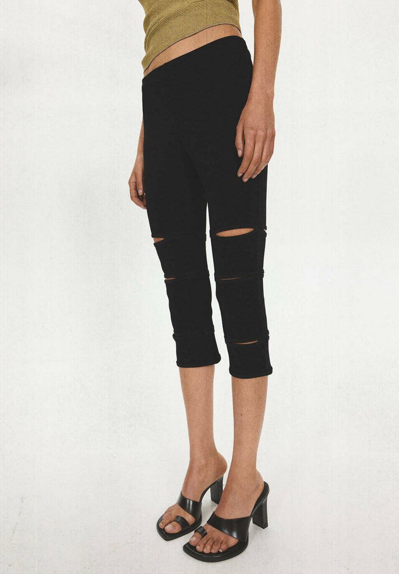 Personne portant un pantalon court noir déchiré et des sandales noires à talons, avec une partie d'un haut sans manches vert visible.