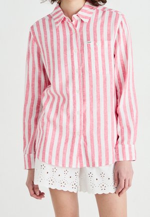 Vrouw draagt een roze-wit verticaal gestreept overhemd met lange mouwen en witte eyelet kanten shorts.