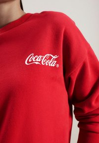 Rode sweatshirt met een wit "Coca-Cola" logo op de linkerkant van de borst. De stof lijkt zacht met een geribbelde halslijn en manchetten.