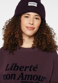 Dark purple knit beanie with a label reading "SENSES THE LABEL" and a matching purple sweatshirt featuring "Liberté mon Amour" in dark text.