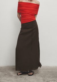 Haut rouge à épaules dénudées, jupe maxi brune taille haute avec fente sur le côté, portée avec des tongs noires. Tissu lisse, silhouette ajustée.