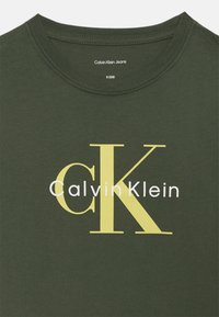 Mørkegrønn bomulls t-skjorte med rund hals som har en stor gul "CK" logo og "Calvin Klein" tekst i hvitt under.