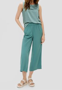 Lichtgroene mouwloze top gecombineerd met losse, wijdvallende teal broeken en crèmekleurige, gevlochten sandalen. Soepele textuur en comfortabele pasvorm.