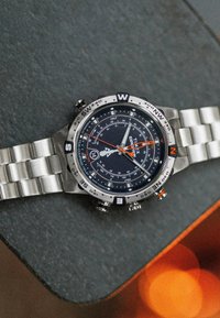 Montre en acier inoxydable argenté avec un cadran bleu foncé, des accents orange, des marquages de boussole et un bracelet à maillons multiple. Elle dispose de trois sous-cadrans.