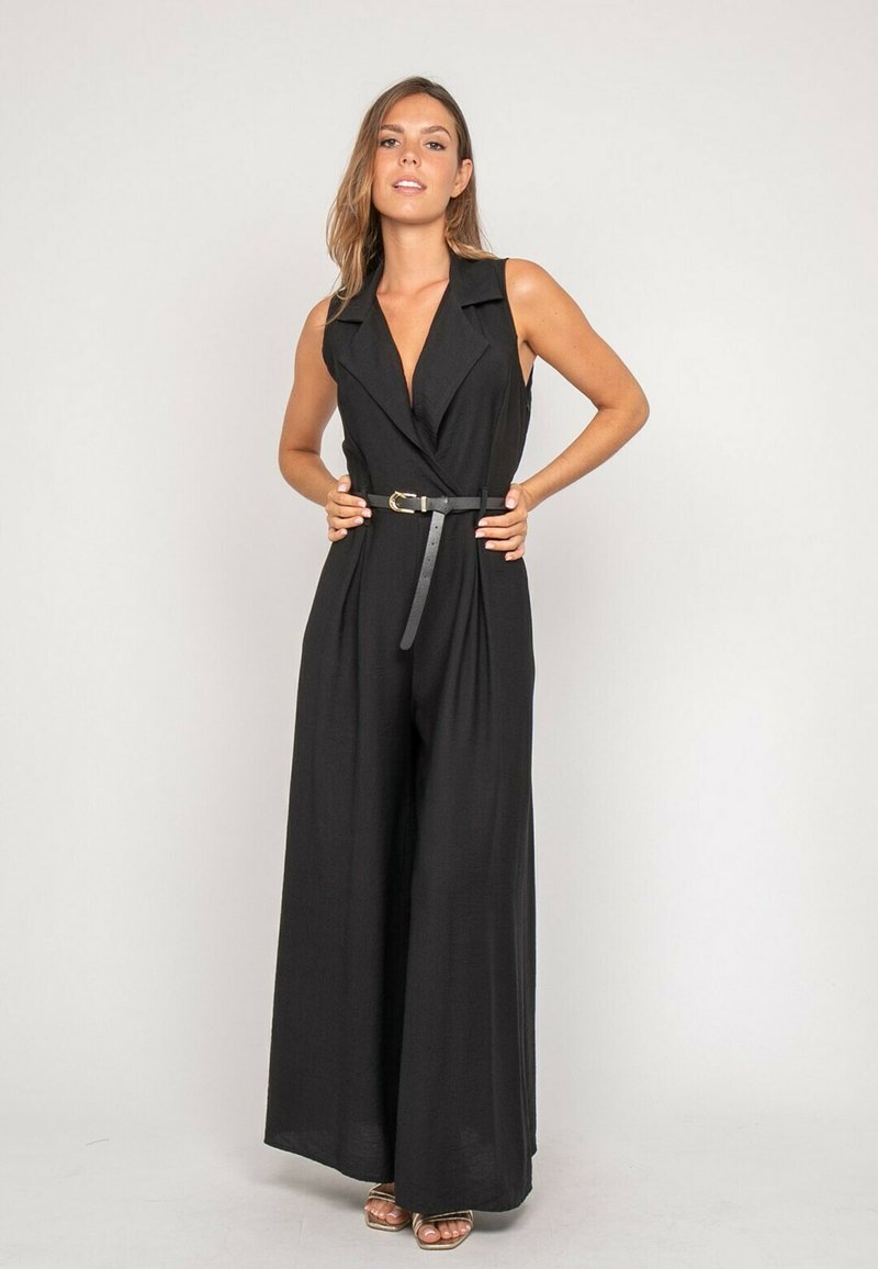 Femme portant une combinaison noire sans manches à jambes larges avec col et ceinture noire, debout les mains sur les hanches devant un fond uni.