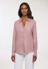 Camicia rosa a maniche lunghe con bottoni e colletto, realizzata in tessuto morbido. Presenta una linea dritta e una leggera lucentezza. Indossata con pantaloni bianchi.
